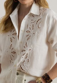 Nő fehér, gombos, virágmintás kivágásos hímzéssel díszített ingben, fehér nadrággal és teknősbéka mintájú, arany karláncokkal.