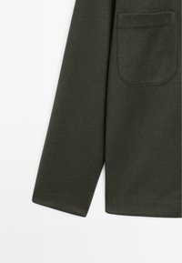 Camicia verde scuro a maniche lunghe con taschino sul petto, vista di lato su sfondo bianco.