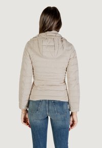 Giubbotto imbottito beige chiaro con cappuccio, caratterizzato da una texture trapuntata, design aderente e una leggera curvatura in vita. Indossato con jeans blu.