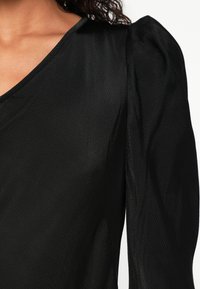 Žena v čiernej blúzke s dlhým rukávom s nadýchanými ramenami a mierne textúrovanou látkou na bielom pozadí.