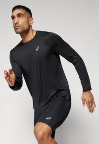 Sort langærmet atletisk skjorte med rund halsudskæring, parret med sorte tætsiddende shorts. Begge beklædningsgenstande har et subtilt logo. Glat tekstur.