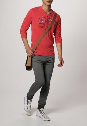 Chemise rouge à manches longues avec un col henley et un détail imprimé, associée à un jean gris et des baskets blanches, portant un sac messager marron.
