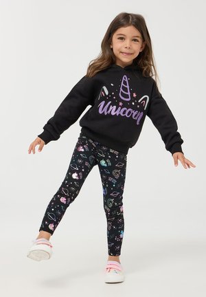Ragazza in equilibrio su una gamba, indossando una felpa nera con il testo "Unicorn" e leggings a fantasia con unicorni e pianeti, sfondo bianco.