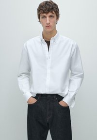 Weißes Baumwollhemd mit Knopfleiste, langen Ärmeln und Kragen. Wird mit dunkelblauer Denimjeans getragen. Schlichtes Design, figurbetonte Passform.