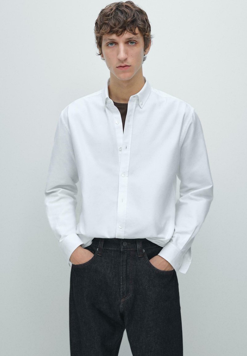 Weißes Baumwollhemd mit Knopfleiste, langen Ärmeln und Kragen. Wird mit dunkelblauer Denimjeans getragen. Schlichtes Design, figurbetonte Passform.