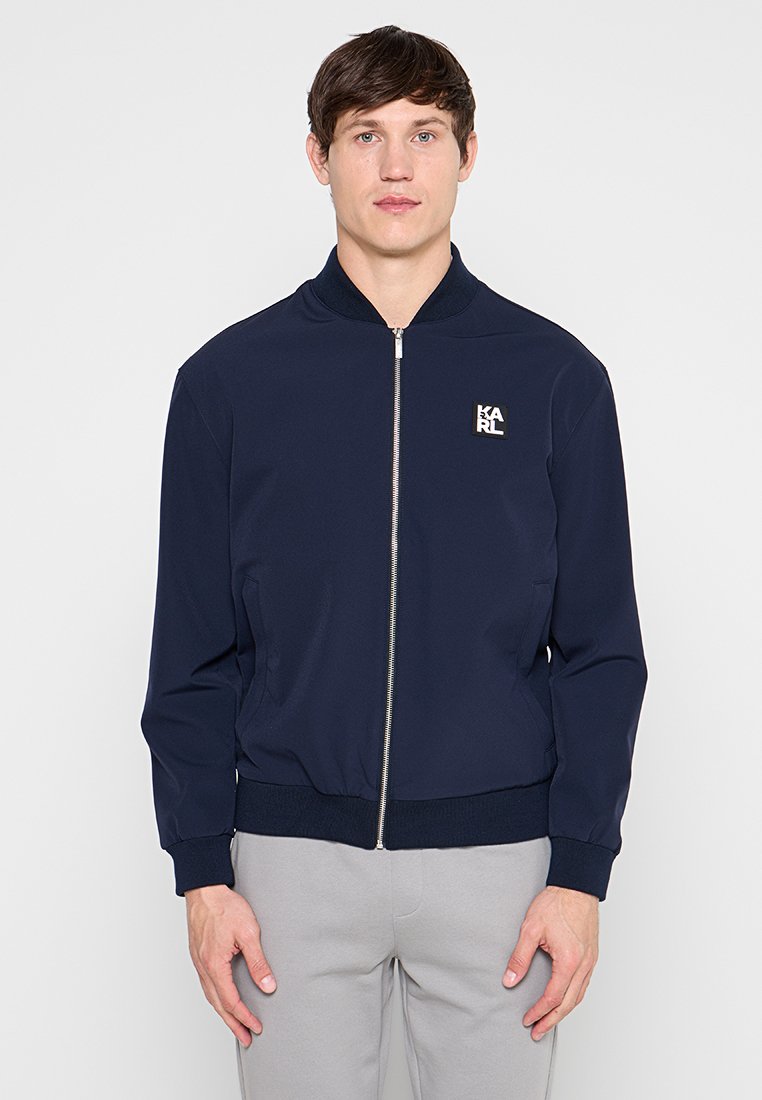 Joven con una chaqueta azul marino con cremallera y un logo "KARL" en el pecho y pantalones gris claro, de pie frente a un fondo blanco liso.