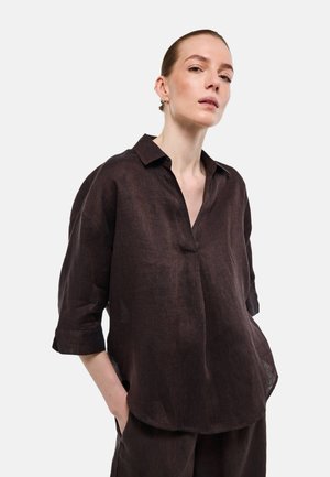Femme portant une chemise ample en lin brun foncé à manches trois-quarts, mains dans les poches, regardant légèrement vers le haut sur un fond uni.
