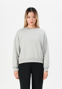 Ung kvinna med långt blont hår bär en ljusgrå sweatshirt och svarta byxor, står mot en helt vit bakgrund.