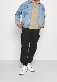 Chaqueta vaquera azul con lavado claro, sudadera con capucha beige, pantalones cargo negros y zapatillas blancas con detalles dorados.