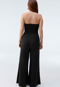 Black strapless jumpsuit with a ruched bodice, wide legs, and smooth fabric. The design features an elastic waistband and flowing silhouette.