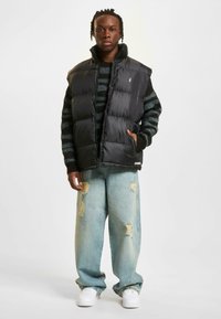 Sort puffer vest med høj krave over en stribet sweater. Båret med slidte lyseblå jeans og hvide sneakers. Subtile logodetaljer.