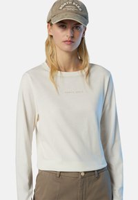 Camicetta a maniche lunghe color crema chiaro con collo rotondo e la scritta "NORTH SAILS" stampata in beige. Abbinata a pantaloni marroni chiari.