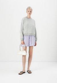 Sweat-shirt gris, shorts rayés bleu et rose, sac à main blanc avec accastillage en argent, tongs noires, fond uni.