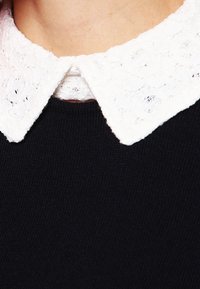 Svart stickad tröja med en vit spetskrage med blommönster och vågiga kanter, vilket visar texturkontrast och lagerdesign.