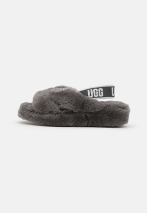 Miękki szary pluszowy kapcie z krzyżującymi się przednimi paskami i elastycznym tylnym paskiem z logo UGG, pokazane na jednolitym białym tle.