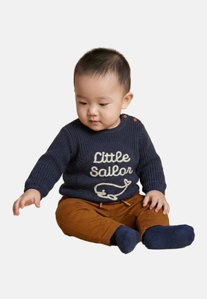 Tout-petit assis, portant un pull bleu marine avec l’inscription « Petit Marin » et un motif de baleine, pantalon marron et chaussettes bleu marine, tendant la main gauche.