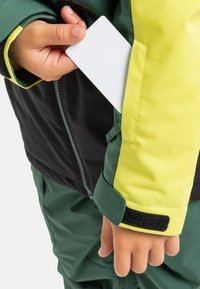 Ein Kind in einer grünen und gelben Jacke steckt eine weiße Karte in die Brusttasche der Jacke.