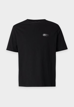 Mand står med front mod kamera iført en sort kortærmet t-shirt, sorte vidde bukser og sorte sko mod en ensartet lys baggrund.
