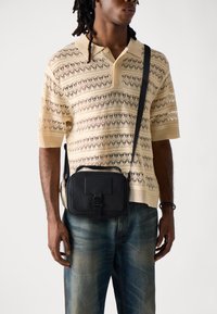 Black crossbody bag with textured surface, rectangular shape, front flap with strap closure, worn over a beige knitted polo shirt.