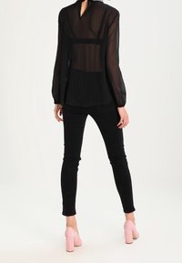 Kvinna iklädd en genomskinlig svart plisserad blus, svarta skinny jeans och rosa högklackade skor, stående med ryggen mot kameran mot en vit bakgrund.