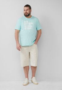 Lichtblauw graphic T-shirt met witte tekst, gecombineerd met crèmekleurige shorts. Het model draagt witte sneakers. Eenvoudige, casual strandlook.