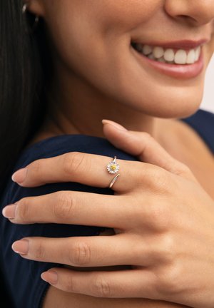 Persona sonriente con piel limpia muestra mano con anillo de plata con diseño de pequeña flor de margarita sobre fondo de tela azul oscuro.