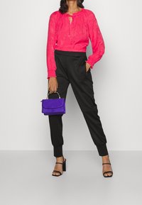 Helles pinkes, langärmliges Bluse mit einem Krawatten-Halsauschnitt und floralem Muster, kombiniert mit schwarzen schmalen Hosen. Hält eine kleine lila Handtasche.