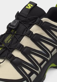 Μπεζ και μαύρο παπούτσι για ορεινό τρέξιμο με σύστημα γρήγορου δέσιμου κορδονιών, υφασμάτινη υφή και μαύρες λεπτομέρειες στη σόλα και το πάνω μέρος.