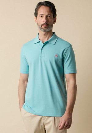 Uomo con capelli corti e scuri e barba che indossa una polo azzurra chiara e pantaloni beige, in piedi davanti a uno sfondo beige.