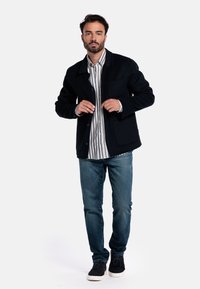 Veste en laine bleu marine avec un col, une fermeture à boutons sur le devant et deux poches avant, superposée sur une chemise rayée blanche et bleu marine, assortie à un jean bleu.