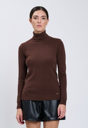 Vrouw met donker haar in een bruine coltrui en zwarte leren shorts, staand tegen een effen lichte achtergrond.