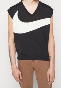 Haut noir sans manches avec décolleté en V et grand motif en forme de swoosh blanc sur la poitrine, porté avec un pantalon marron par une personne ayant des tatouages visibles sur les bras.