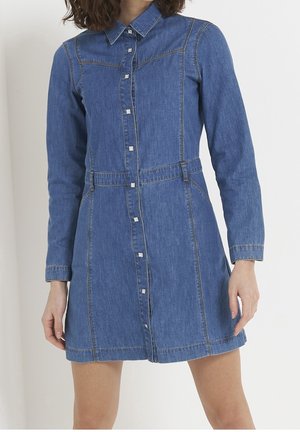 Kobieta ubrana w niebieską dżinsową sukienkę zapinaną na guziki z długim rękawem, kołnierzykiem i widocznym przeszyciem, stojąca na jednolitym białym tle.