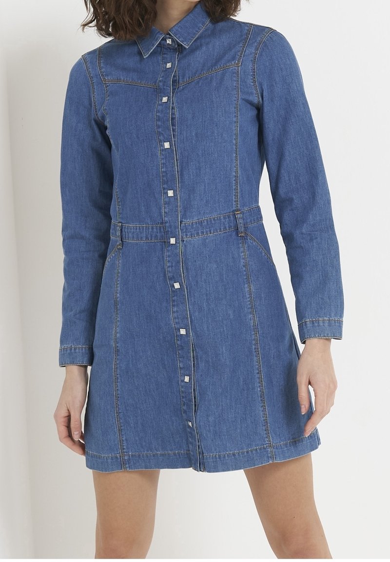 Mujer vistiendo un vestido azul de mezclilla con botones largos, cuello y costuras visibles, de pie frente a un fondo blanco liso.