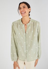 Blusa de botones verde claro con cuello rizado, con bordado de hojas blancas y mangas largas, confeccionada en una tela suave y con textura.