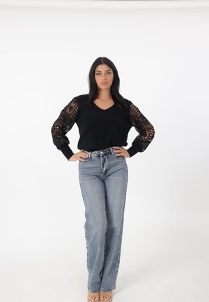 Donna in piedi con le mani sui fianchi, indossa un maglione nero con maniche in pizzo e jeans a vita alta azzurri su sfondo bianco.