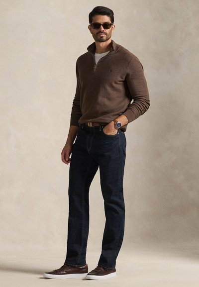 Rudas megztinis su užtrauktuku, tamsiai mėlyni džinsai, rudi sportbačiai ir akiniai nuo saulės. Išskirtinės savybės: briaunotas raštas, laikrodis ir diržas. Neutralus fonas.