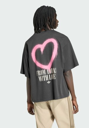 Person mit Dreadlocks, die ein schwarzes oversized T-Shirt mit einem pink gesprayten Herz und dem Schriftzug "FROM ADIDAS WITH LOVE" auf dem Rücken trägt.