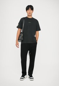 Schwarzes T-Shirt mit grünem Logo, kombiniert mit schwarzen, schmal zulaufenden Hosen und Sneakers mit weißen Akzenten. Dazu gehört auch eine kleine schwarze Umhängetasche.