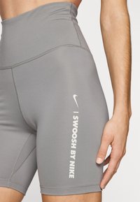 Pantaloni corti grigi a vita alta realizzati in tessuto elasticizzato, con logo Nike e testo "I SWOOSH BY NIKE" in bianco lungo il lato.