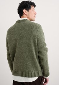 Maglione verde in maglia testurizzata con orlo e polsini a coste. Presenta un colletto alto e viene indossato sopra una camicia bianca. Vestibilità casual.