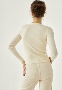 Suéter de crema con diseño ajustado y acanalado, mangas largas y cuello redondo, combinado con pantalones beige claros que tienen bolsillos traseros.