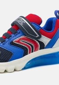 Baskets de sport bleues et rouges avec des détails en maille, une sangle velcro, une semelle en caoutchouc et une section de talon bleu transparent. Présente un accent de design avec des rayures.