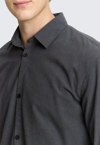 Dunkelgraues Hemd mit Knopfleiste und Kragen, aus texturiertem Stoff mit schwarzen Knöpfen, zeigt eine glatte Oberfläche und einen dezenten Glanz.