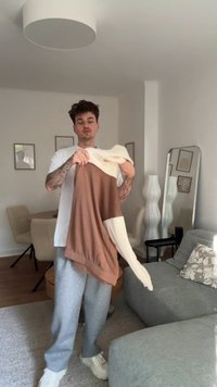 Maglione marrone e crema con maniche lunghe e una vestibilità rilassata, tenuto con una mano. Indossato con pantaloni della tuta grigi e scarpe da ginnastica bianche in una stanza luminosa.