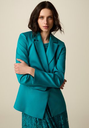 Donna con capelli scuri ondulati che indossa un blazer turchese sopra un vestito di pizzo, in piedi con le braccia incrociate davanti a uno sfondo beige uniforme.