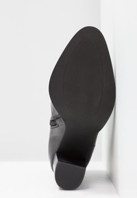 Semelle de bottine en cuir noir avec un petit talon, montrant une bande de roulement texturée, positionnée verticalement contre une surface blanche en angle.