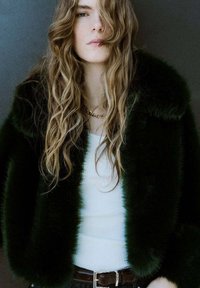 Veste en fausse fourrure verte à la texture moelleuse, avec un large col. Portée par-dessus un haut blanc côtelé et complétée par un collier chaîne en or.