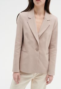 Beige kavaj med slagkrage, enkel knappstängning och sidofickor. Slät, strukturerad tyg ger ett skräddarsytt utseende.