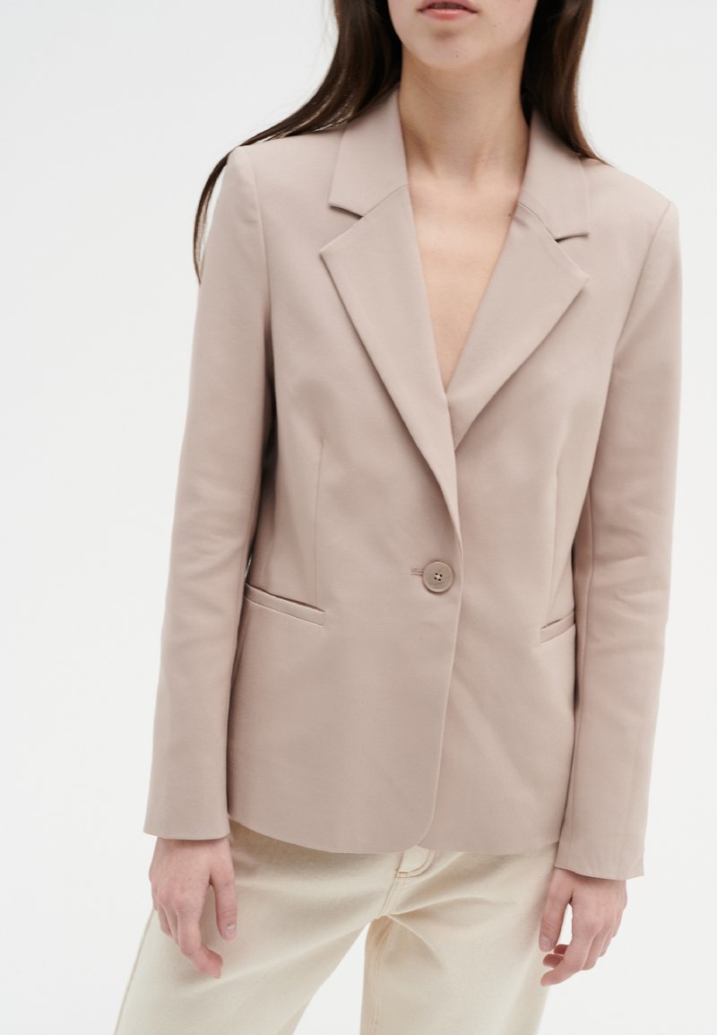 Beige kavaj med slagkrage, enkel knappstängning och sidofickor. Slät, strukturerad tyg ger ett skräddarsytt utseende.